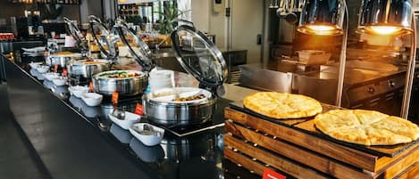 A buffet area with various food containers, a pizza display, and a kitchen area with stainless steel appliances.