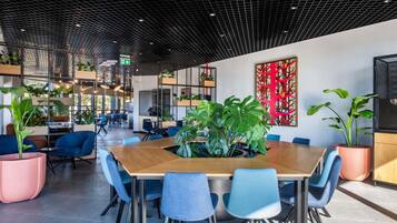 A modern indoor space with a wooden conference table, blue chairs, potted plants, and a grid-patterned ceiling.