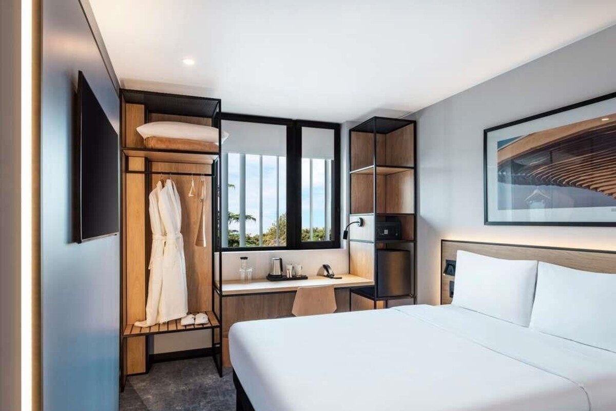 Een moderne hotelkamer met een bed, bureau, stoel, kledingkast en groot raam.