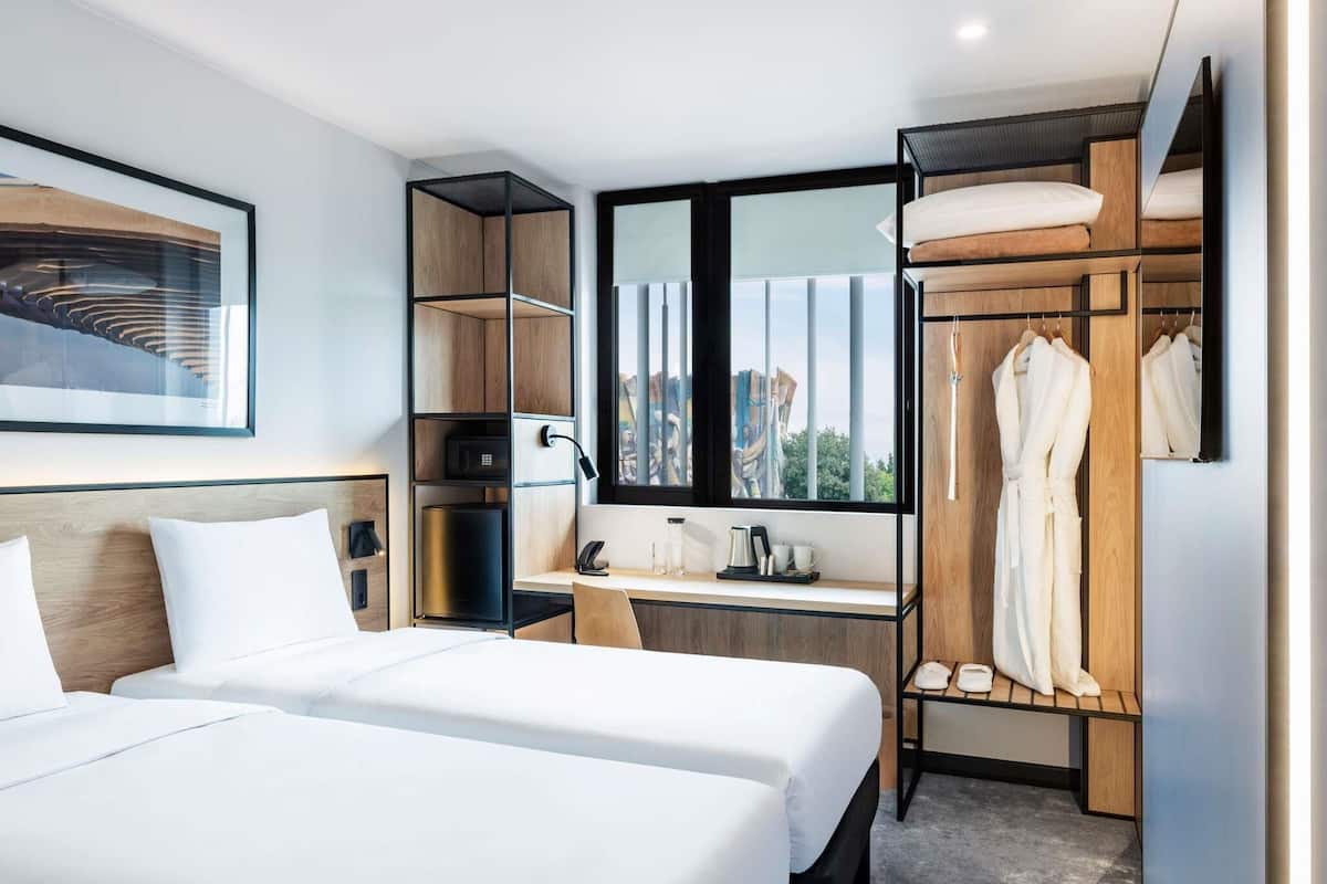 A modern hotel room with two beds, a desk, a wardrobe, and a large window.