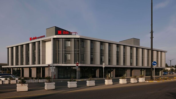 A modern hotel building with a prominent 'ibis hotel' sign, large windows, and a street view with a directional sign. - Ibis Tbilisi Airport (Tbilisi)