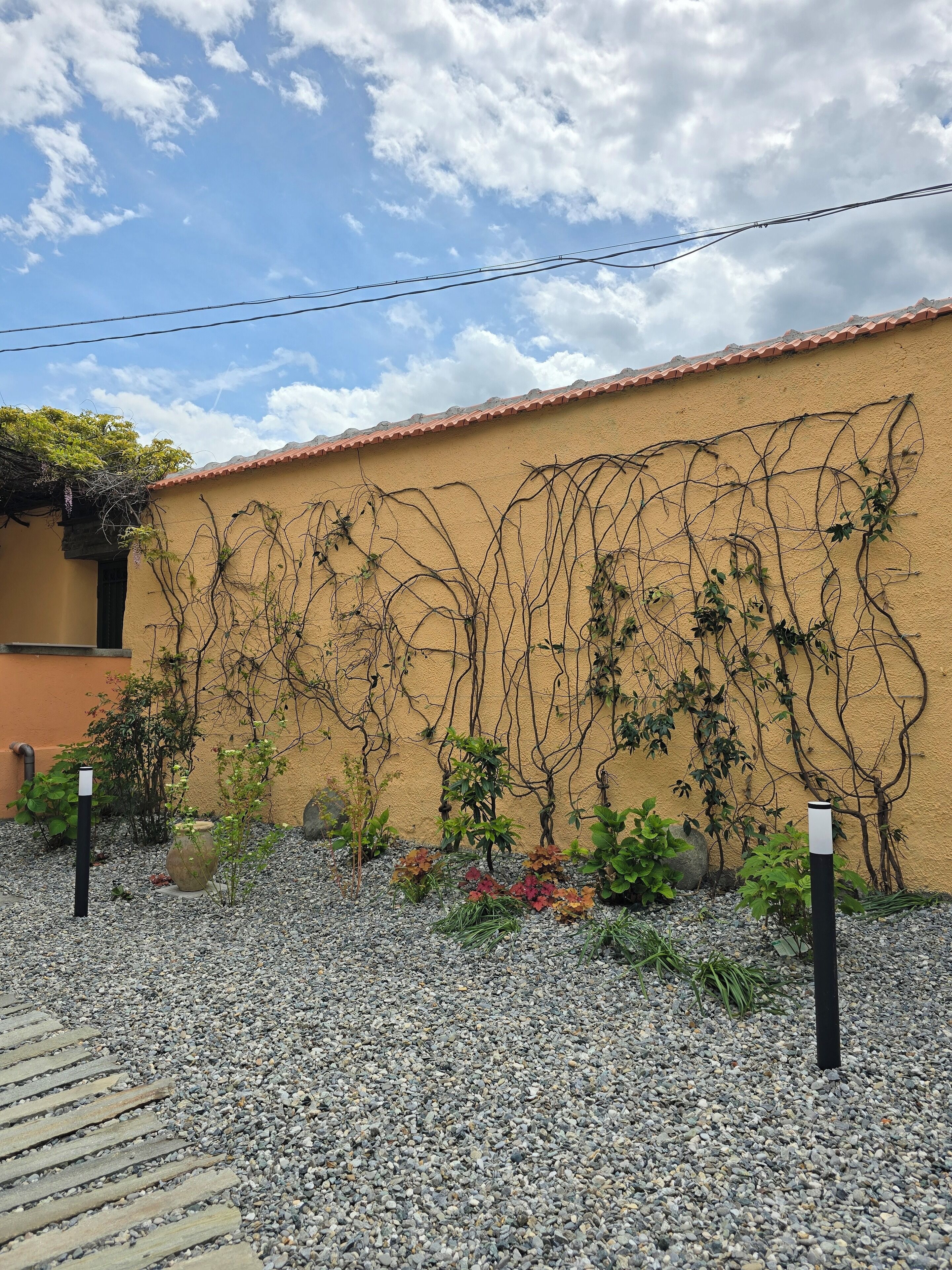 Un edificio con un muro giallo e un tetto in tegole rosse. Davanti all'edificio si estende un giardino con ghiaia e piante.