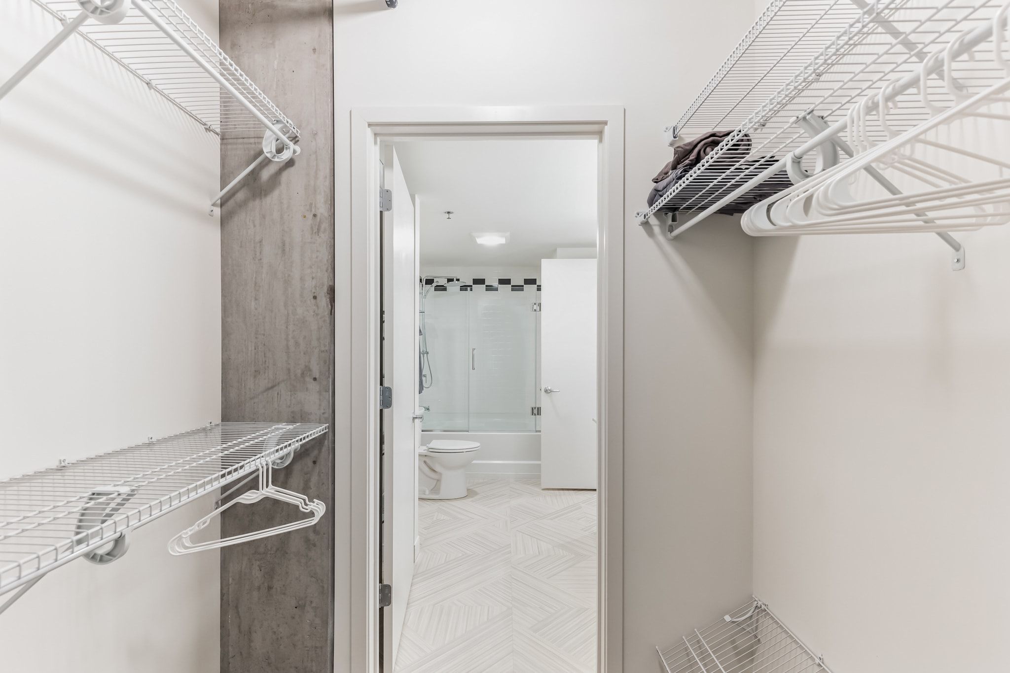 Un baño estrecho con paredes de azulejos blancos, suelo con estampado de espiga y estantes metálicos para tender ropa.