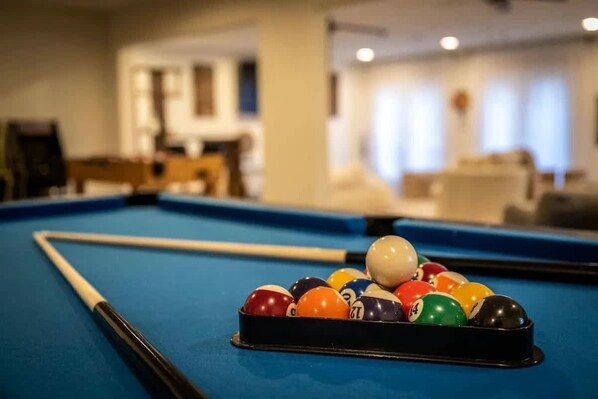 A pool table with a set of billiard balls in the center, a pool cue, and a blurred background suggesting a room with furniture and windows.