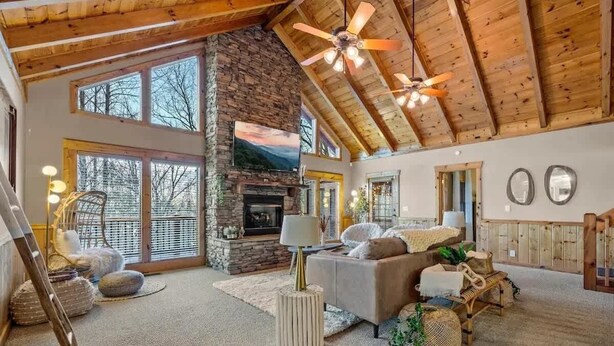 A spacious living room with a stone fireplace, large windows, and a high ceiling with exposed beams. - Round Top Way Cabin by Beyond Expectations: 6BR Luxury Cabin w/ Hot Tub & Sauna (Sevierville)