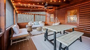 A cozy cabin interior with a wooden ceiling, string lights, a ceiling fan, a wooden table with benches, a cushioned bench with pillows, and a window with a view of trees. - Round Top Way Cabin by Beyond Expectations: 6BR Luxury Cabin w/ Hot Tub & Sauna (Sevierville)