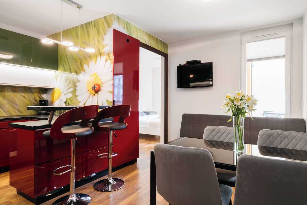 A modern living room with a red kitchen area, a dining table with chairs, and a TV mounted on the wall.
