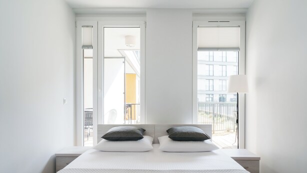 A modern bedroom with a large bed, two pillows, a nightstand, and a window with blinds. - Native Apartments Masarska 8 (Kraków)