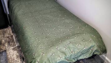 A single bed with a green bedspread featuring a pattern of small, stylized bees. The bed is positioned against a white wall, and there is a wooden floor with a rustic appearance.