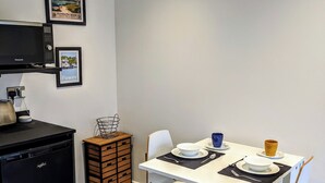 A compact kitchen with a black refrigerator, a microwave, and a small wooden cabinet. A dining table with white chairs and a black placemat is set with a bowl, a cup, and a plate. - The Orchid (Gillingham)