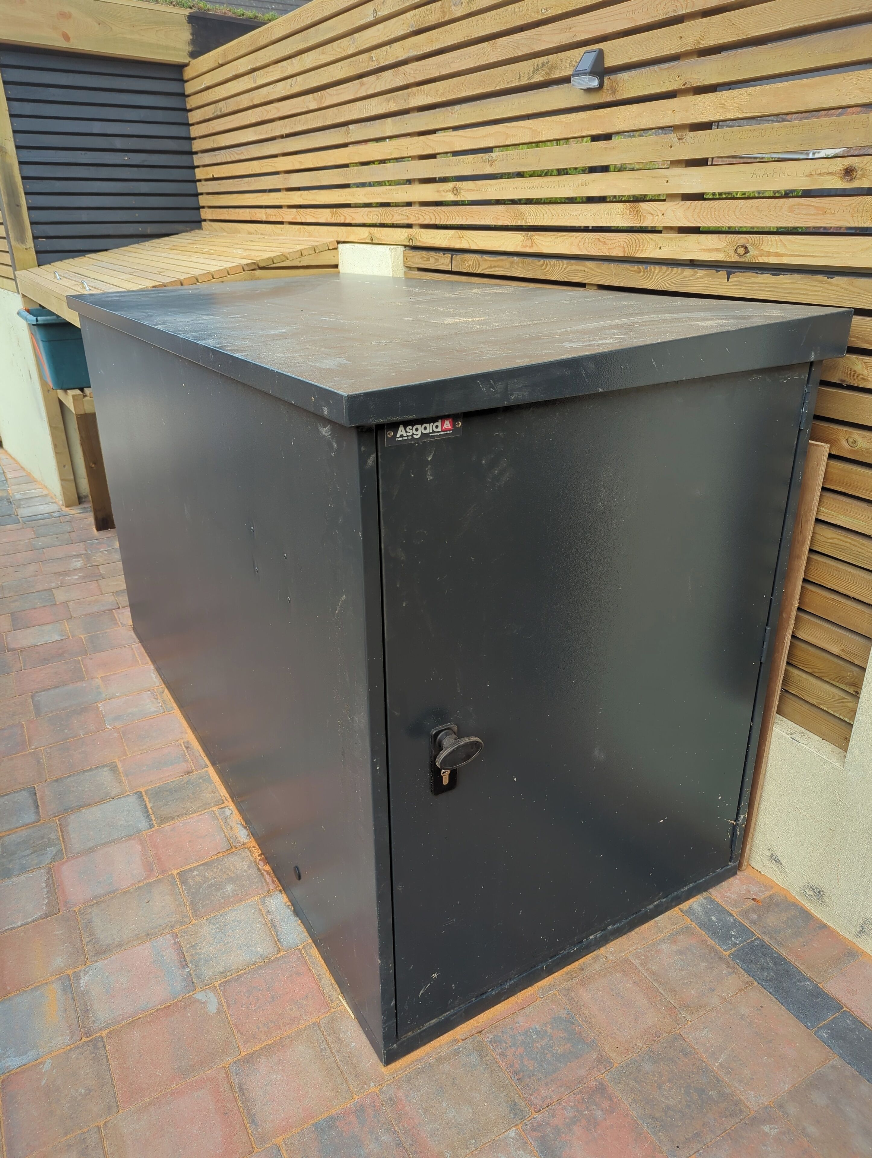 Un mueble de almacenamiento metálico negro con cerradura, situado sobre un área pavimentada con ladrillos junto a una cerca de madera.