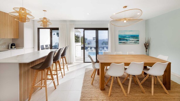 Una cocina moderna con una isla central, un comedor con mesa de madera y sillas blancas, y vista al mar a través de grandes ventanales.