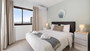 Una habitación de hotel con una cama grande, dos mesitas de noche, un ventanal con cortinas, una obra de arte en la pared y vistas a la ciudad.