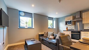 A modern living room with a leather sofa, a coffee table, and a TV. There are windows with plants, a kitchen with a microwave, and a dining area with a table and chairs. - Two Bedroom Newcastle City Quayside Apartment (Newcastle-upon-Tyne)