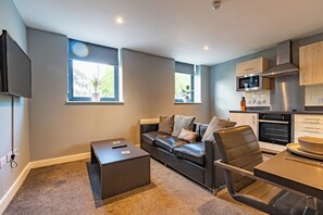 A modern living room with a leather sofa, a coffee table, and a TV. There are windows with plants, a kitchen with a microwave, and a dining area with a table and chairs. - Two Bedroom Newcastle City Quayside Apartment (Newcastle-upon-Tyne)