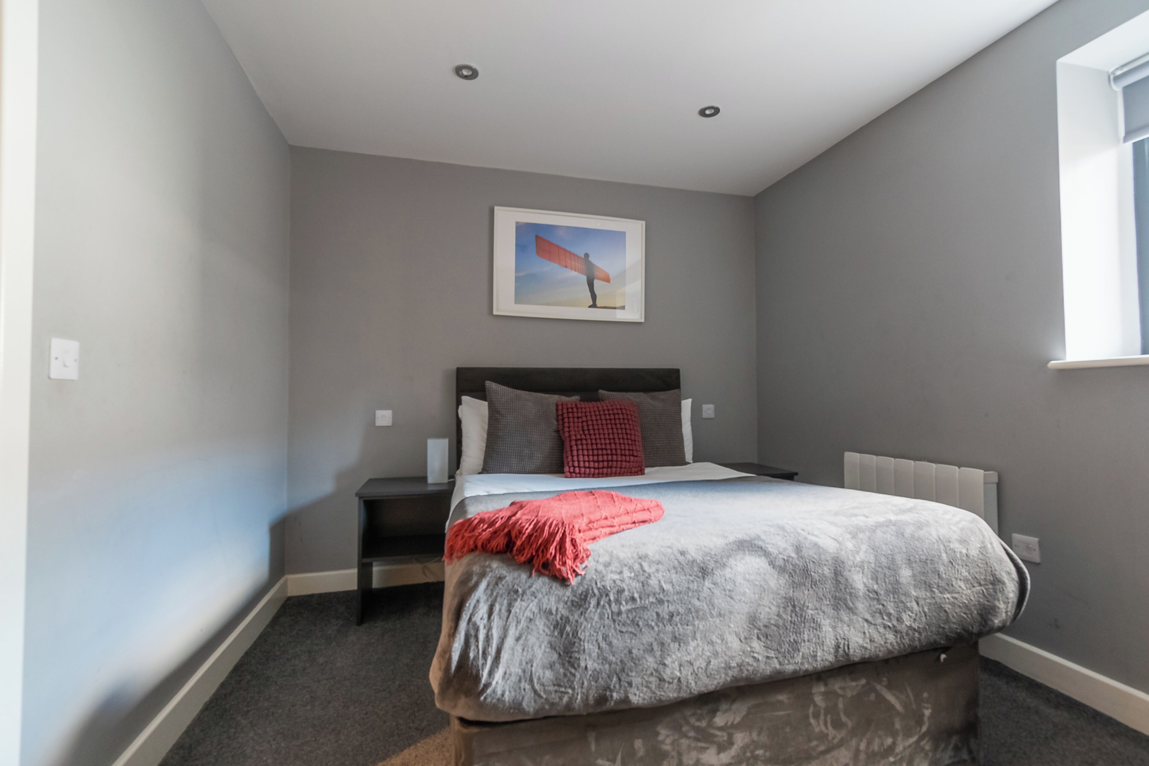 A bedroom with a bed, a nightstand, a red throw blanket, and a framed picture on the wall.