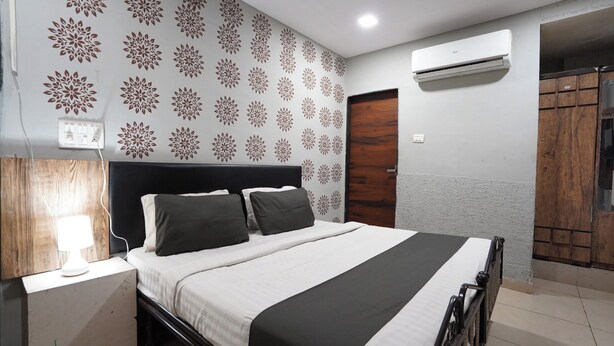 A bedroom with a bed, a bedside table with a lamp, patterned wallpaper, an air conditioning unit, and a wardrobe. - Hotel Sai Inn (Ahmedabad)