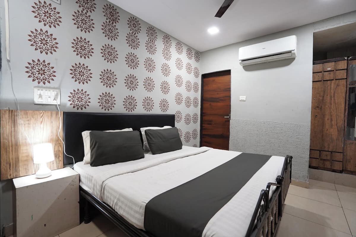 A bedroom with a bed, a bedside table with a lamp, patterned wallpaper, an air conditioning unit, and a wardrobe.