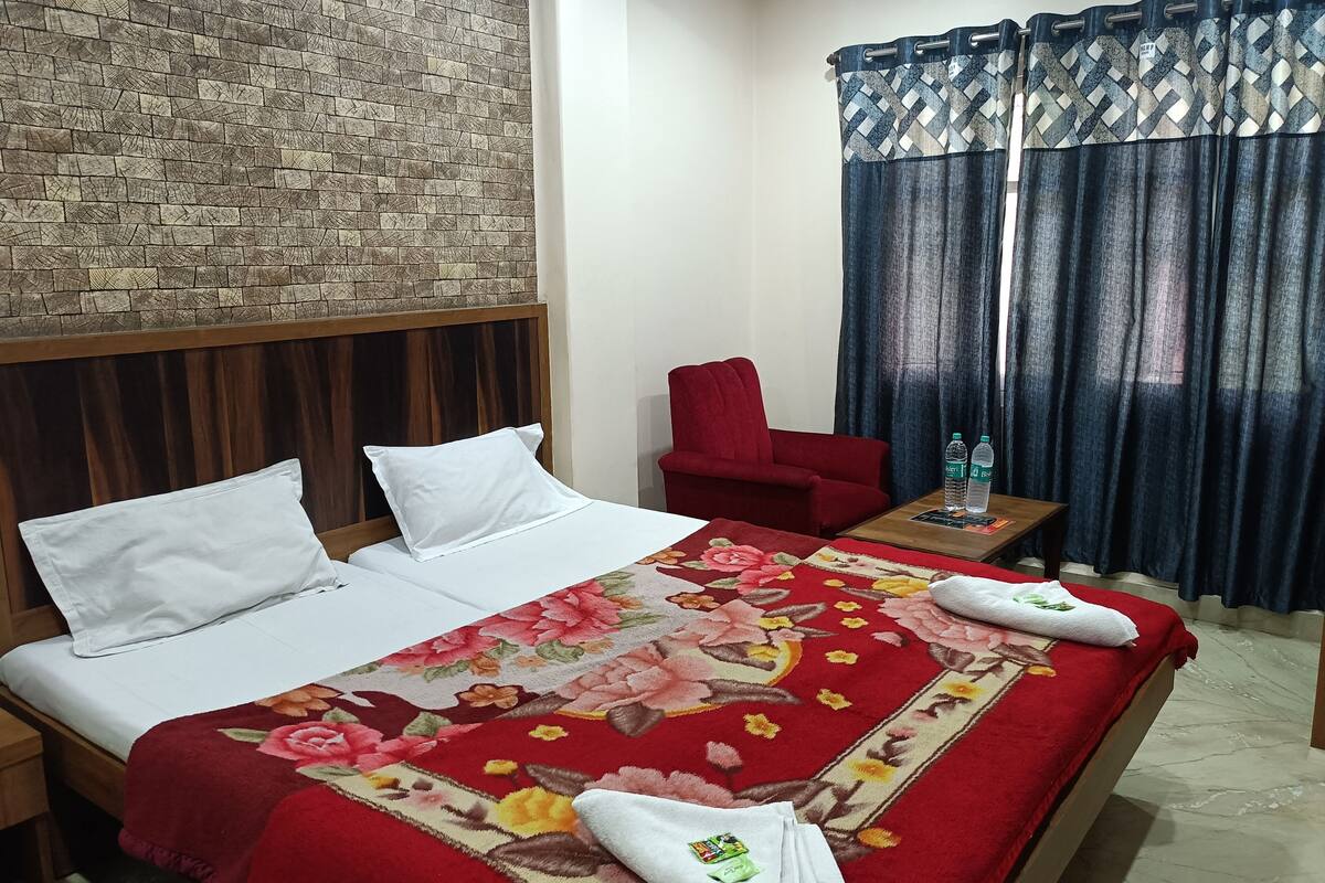 A hotel room with a bed, a red chair, a small table with water bottles, and a window with blue curtains.