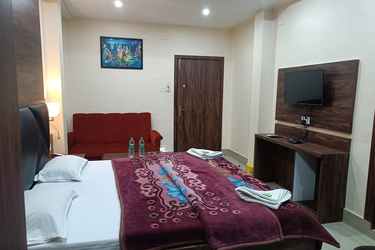 A hotel room with a bed, a red sofa, a wooden wardrobe, a television, and a wall-mounted TV.