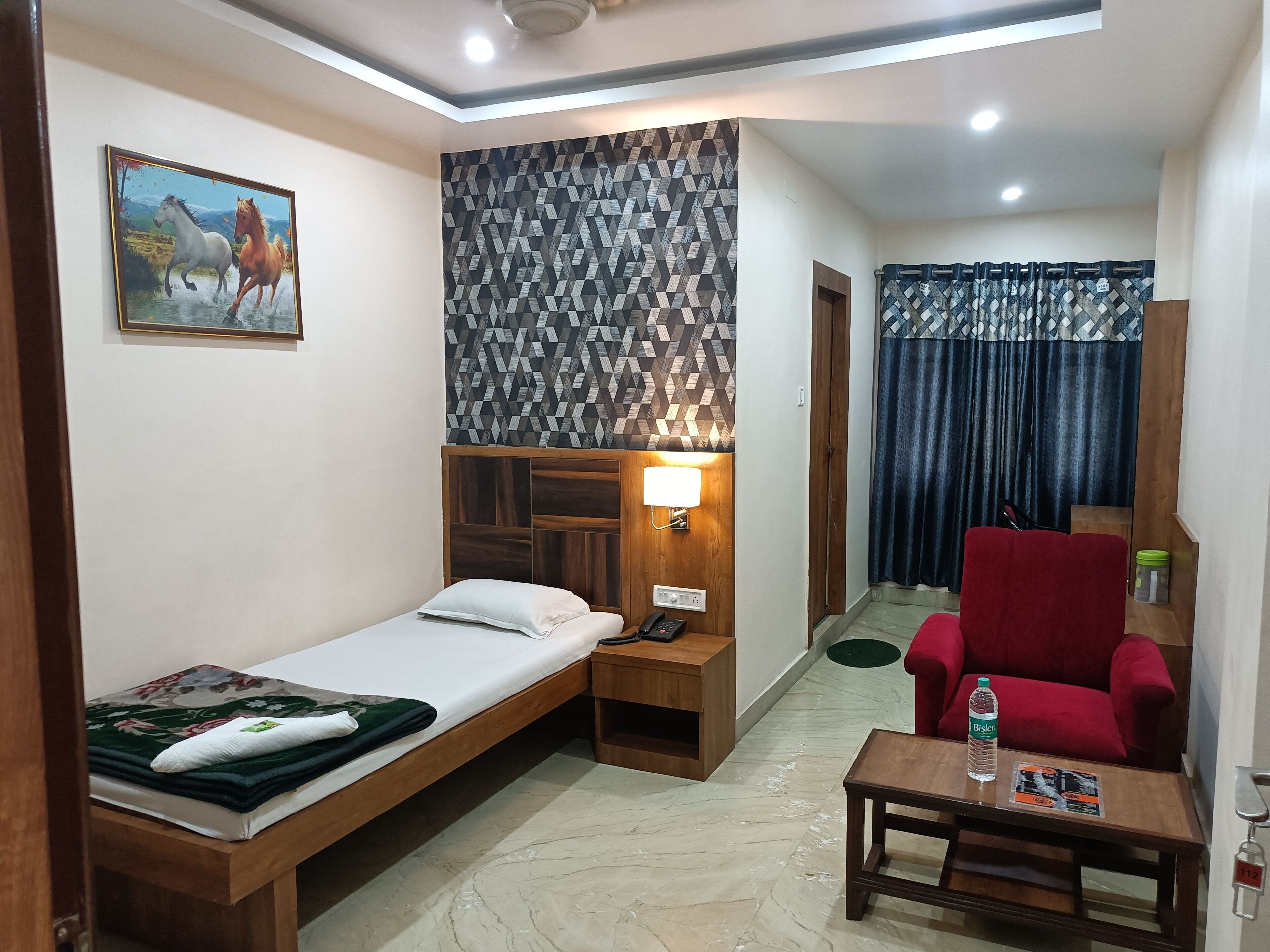 A compact hotel room with a single bed, a wooden nightstand with a lamp, a red armchair, a small table with a water bottle, and a patterned wall.