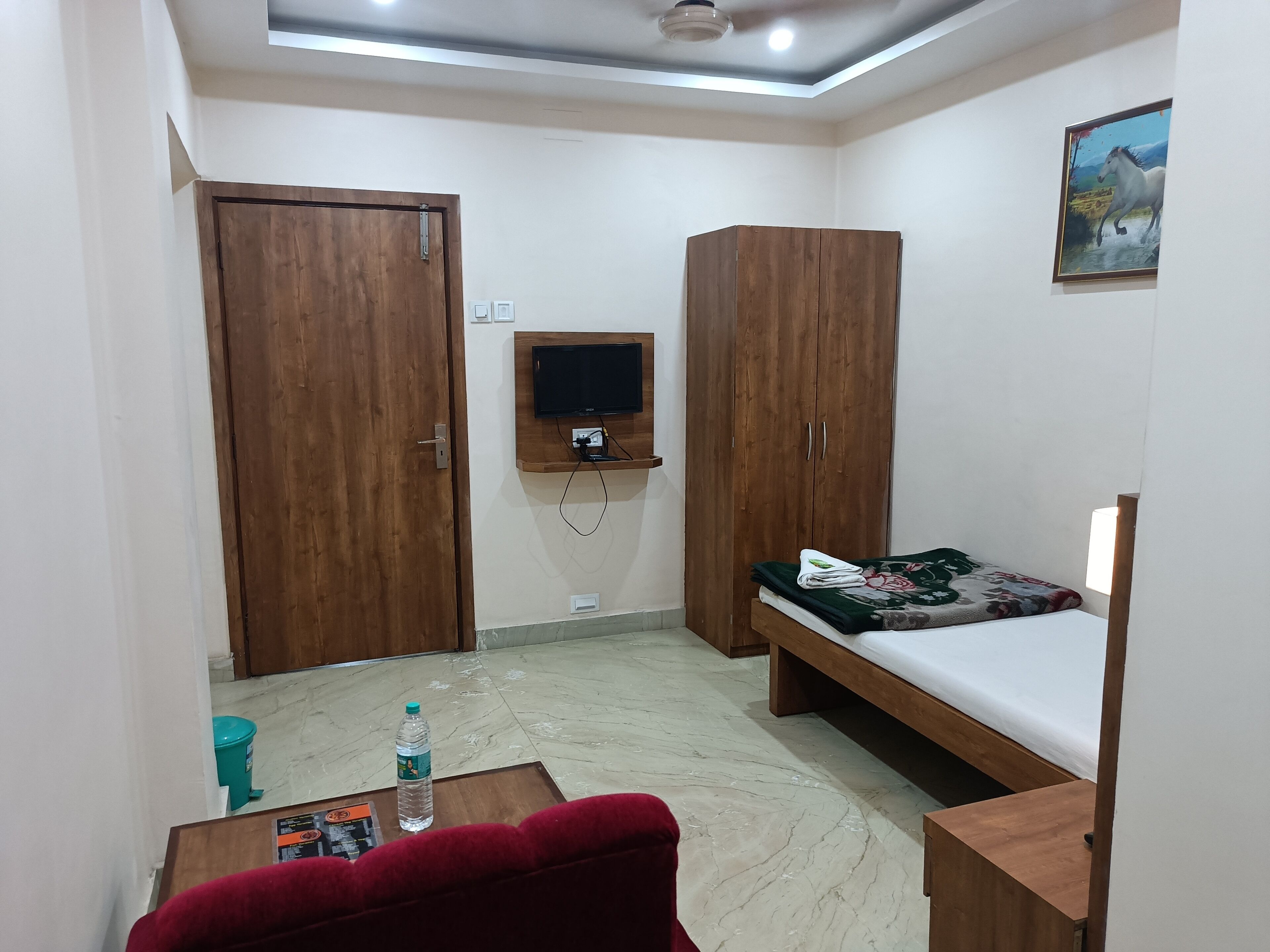 A small, well-lit room with a bed, two wardrobes, a mounted TV, a water bottle, and a magazine on the floor.
