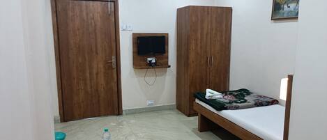 A small, well-lit room with a bed, two wardrobes, a mounted TV, a water bottle, and a magazine on the floor.