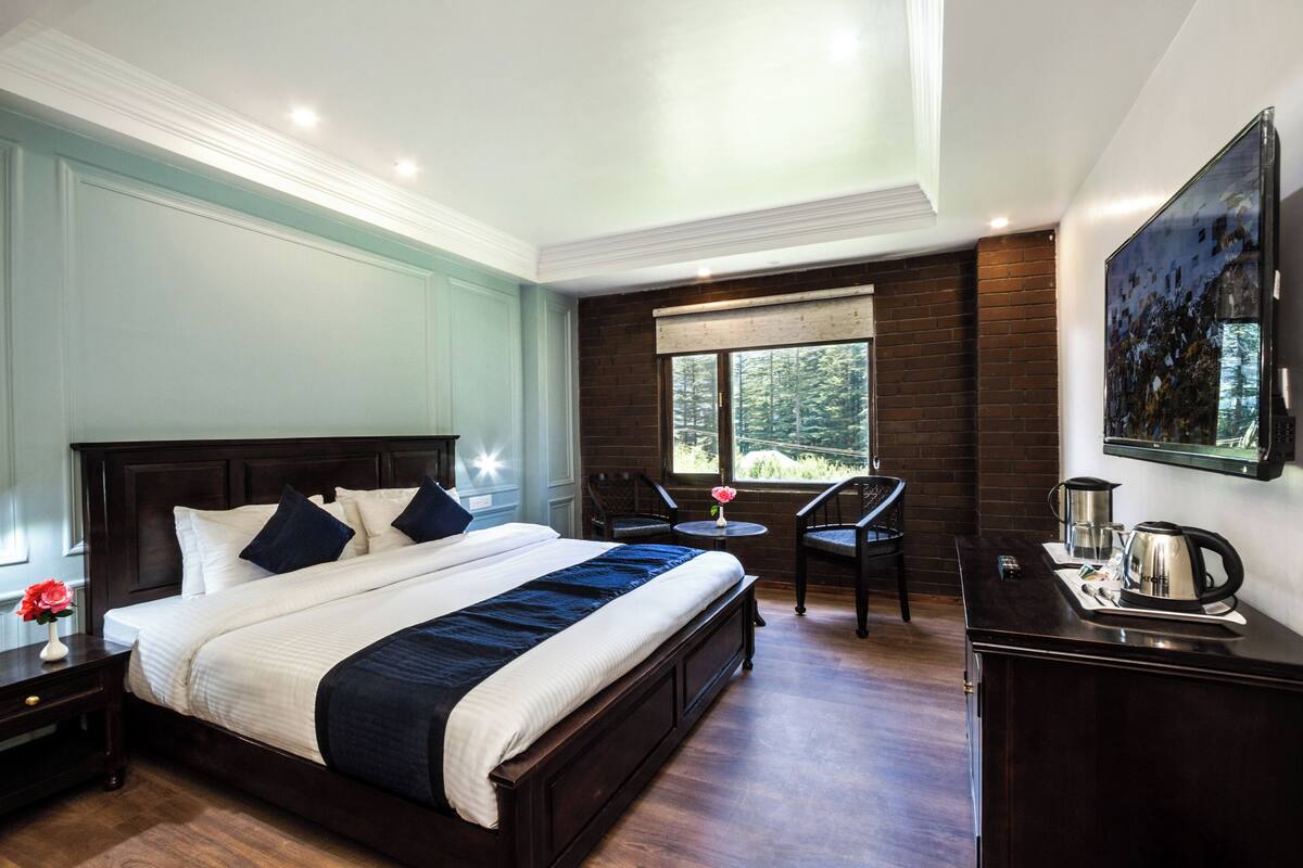A bedroom with a large bed, a small table with chairs, a coffee maker, and a window with a view of trees.