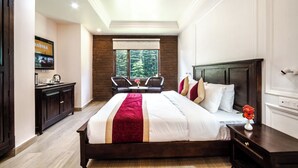 A hotel room with a large bed, a wooden headboard, a nightstand, a TV, a window with a view of trees, and a small table with chairs. - Elysian Woods New Devidayar (Manali)
