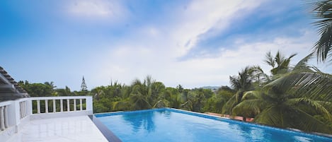 A swimming pool with a clear blue water surface, surrounded by lush greenery and palm trees, with a white tiled deck and a metal ladder leading into the pool.
