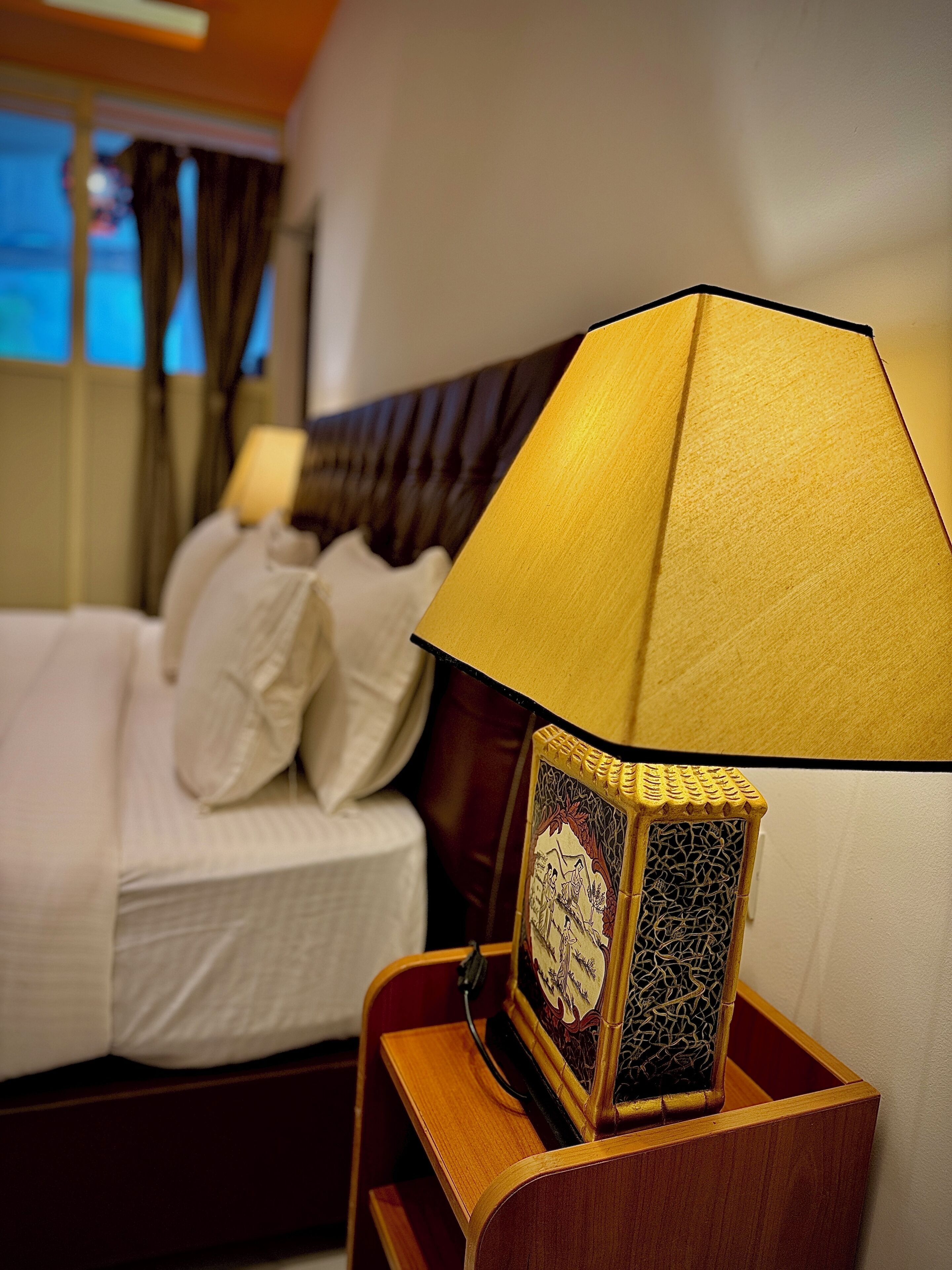 A neatly made bed with white linens and pillows, a wooden bedside table with a lamp and a decorative item, and a window with curtains in the background.