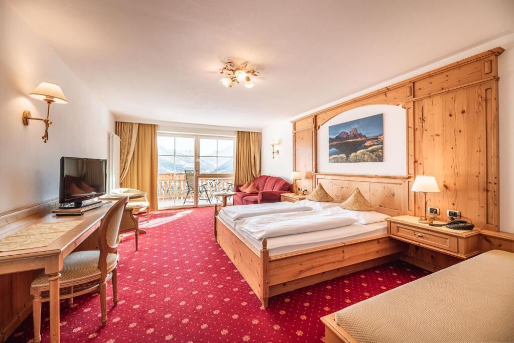 A hotel room with a wooden bed, a desk with a chair, a television, and a balcony with a view of mountains.