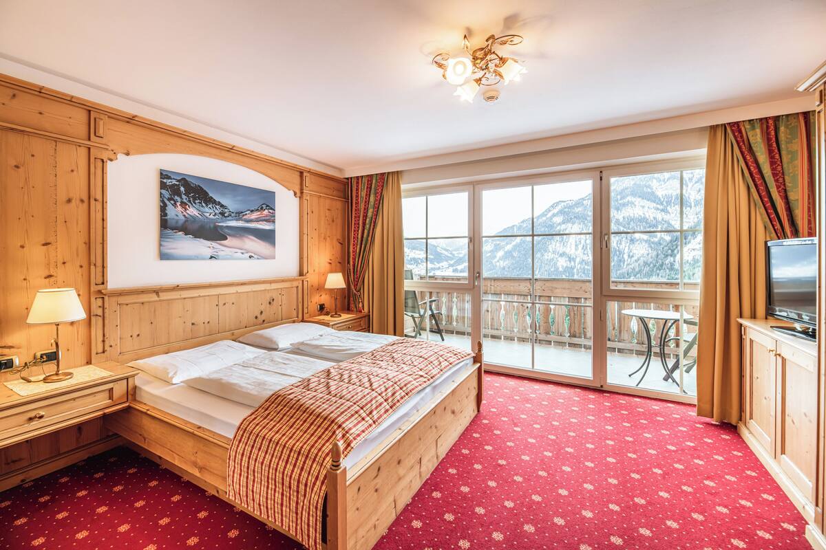 Une chambre d’hôtel avec un lit en bois, une grande fenêtre donnant sur la montagne, un balcon avec une table et des chaises, et un téléviseur posé sur une commode en bois.