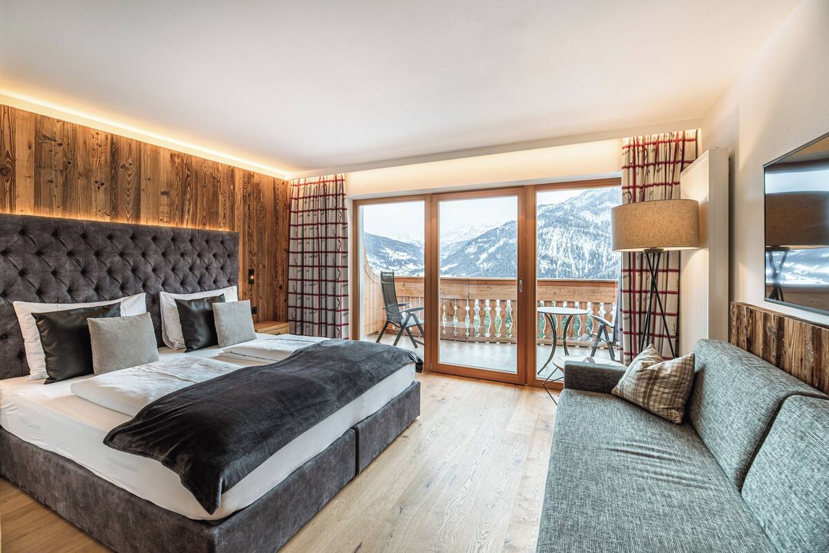 Une chambre dotée d’un grand lit, d’un canapé, d’un balcon offrant une vue sur les montagnes et de lambris en bois sur les murs.