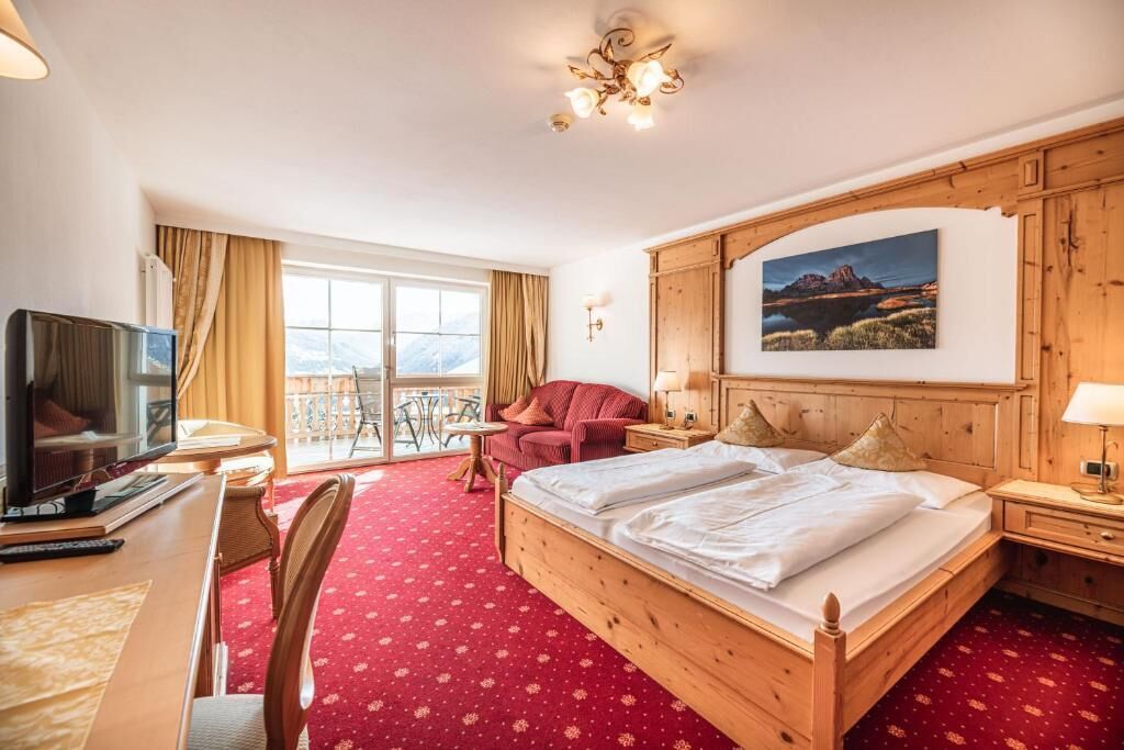 A hotel room with a wooden bed, a desk with a TV, a chair, a balcony with a view, and a painting on the wall.
