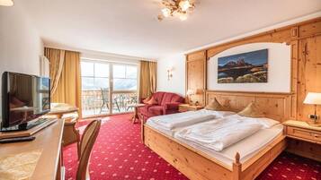 A hotel room with a wooden bed, a desk with a TV, a chair, a balcony with a view, and a painting on the wall.