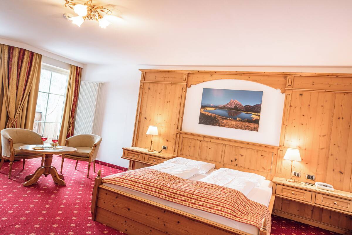 Une chambre d’hôtel avec un lit en bois, un bureau avec une chaise, une lampe et un tableau représentant un paysage de montagne accroché au mur.