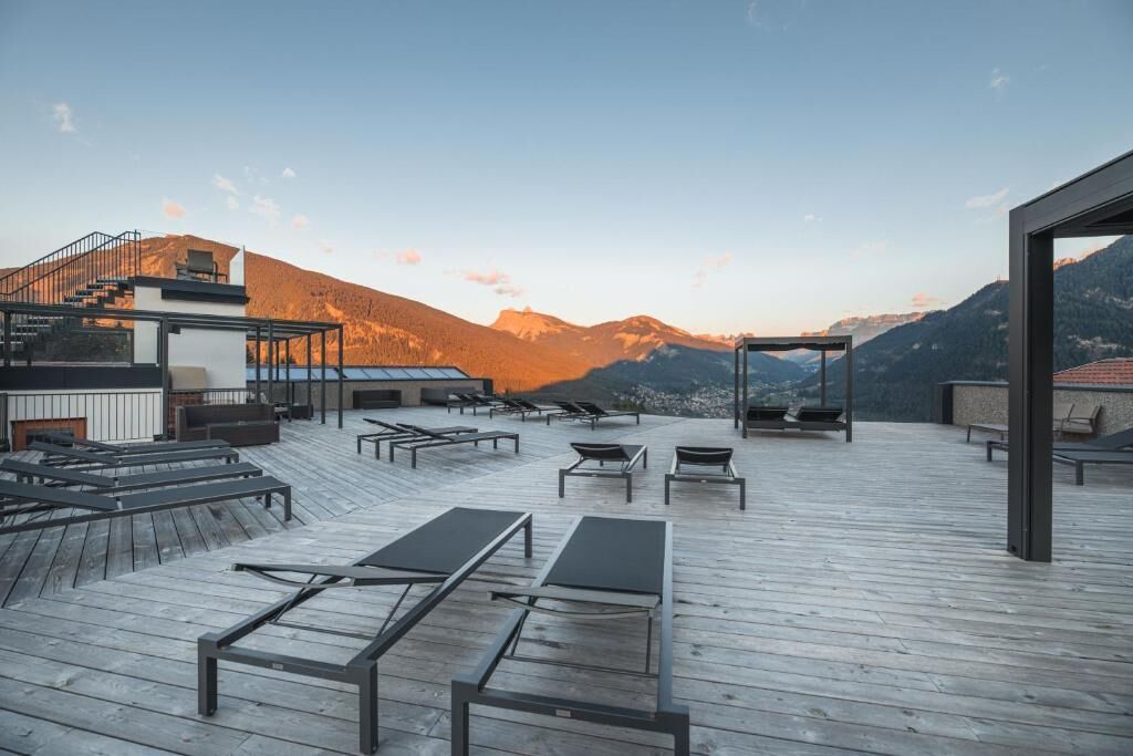 A rooftop terrace with wooden flooring, sun loungers, and a view of mountains.