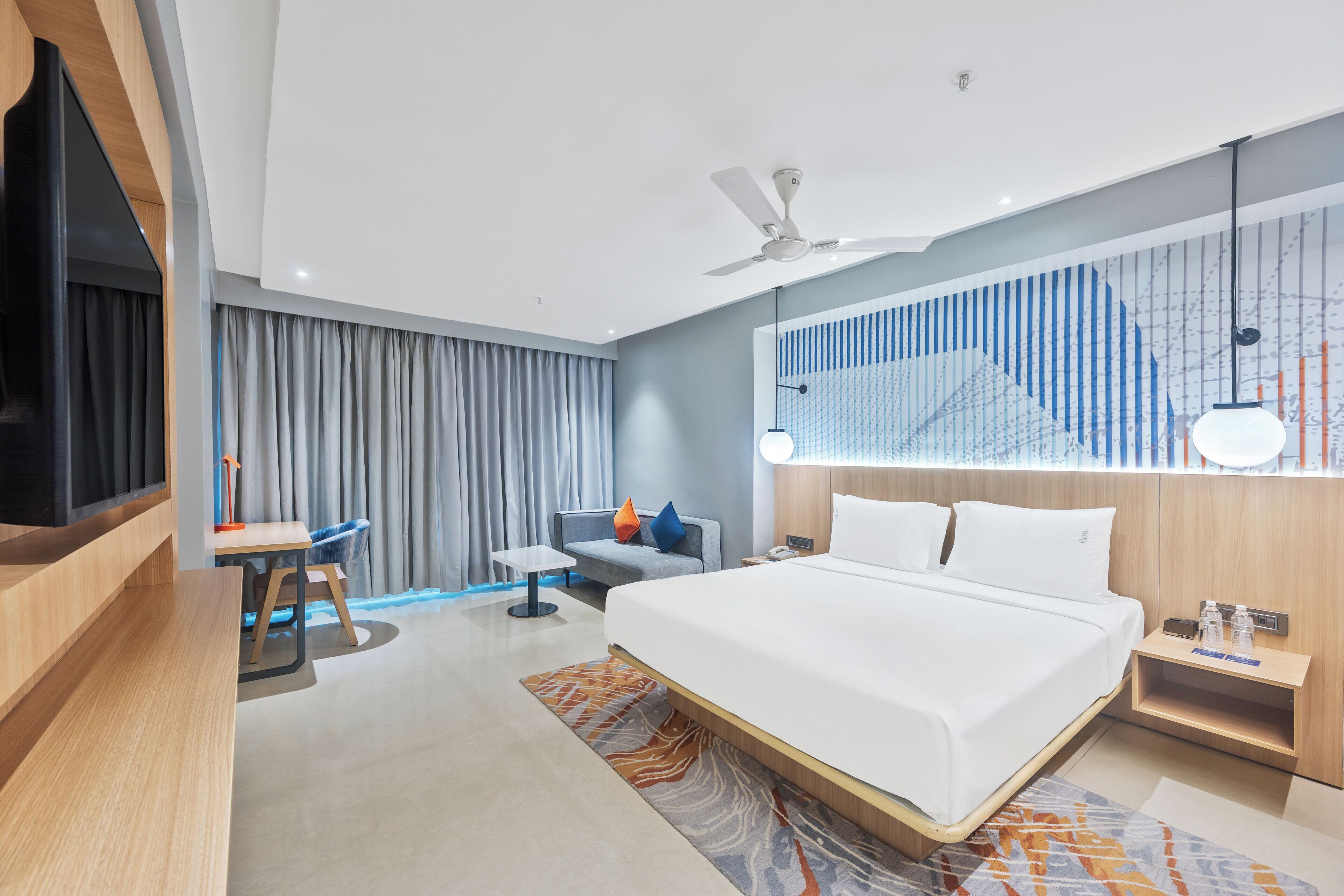 Ein modernes Hotelzimmer mit einem großen Bett, einem Schreibtisch, einem Sessel, einem Fernseher, einem Deckenventilator und einem großen Fenster mit Vorhängen.