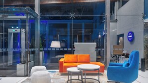 Een moderne lobby met een glazen lift, een oranje bank, een witte poef, een blauwe stoel en een kleine tafel.