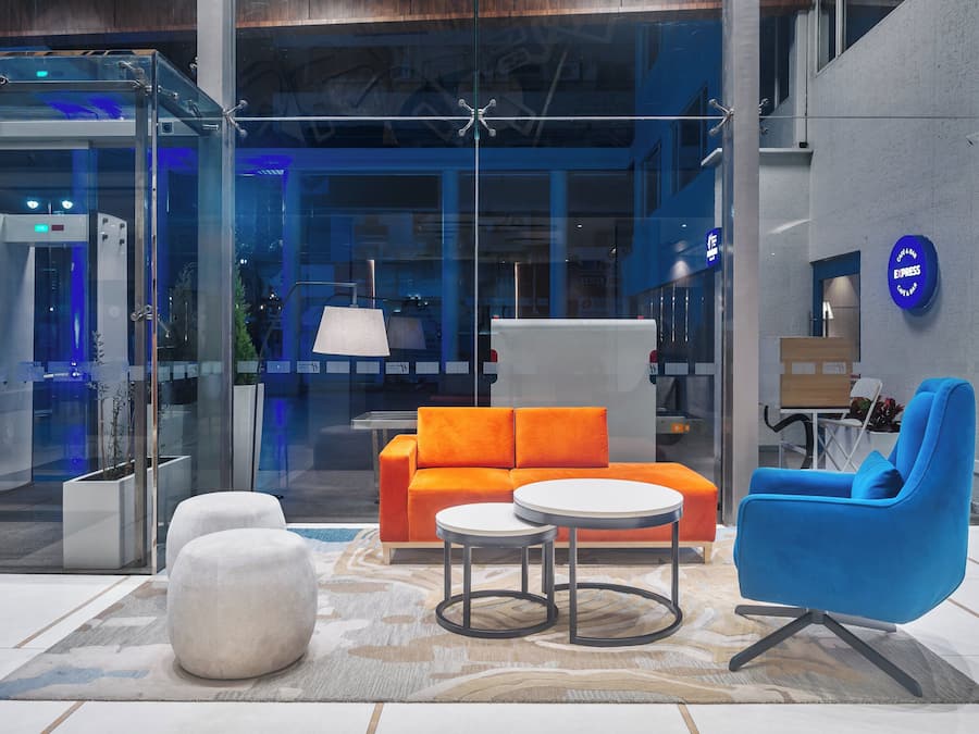 Een moderne lobby met een glazen lift, een oranje bank, een witte poef, een blauwe stoel en een kleine tafel.