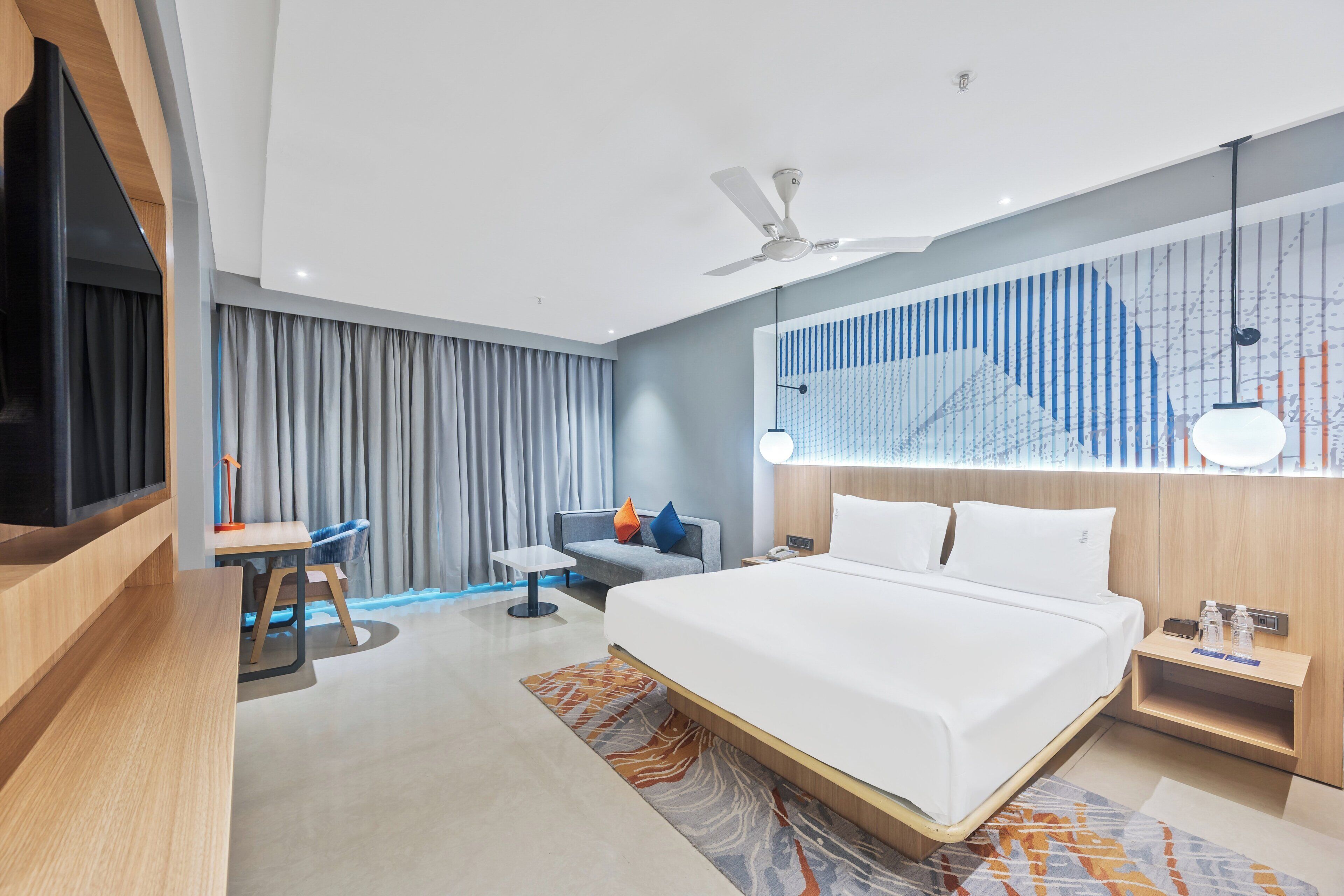 A modern hotel room with a large bed, a desk, a chair, a ceiling fan, and a large window with curtains.