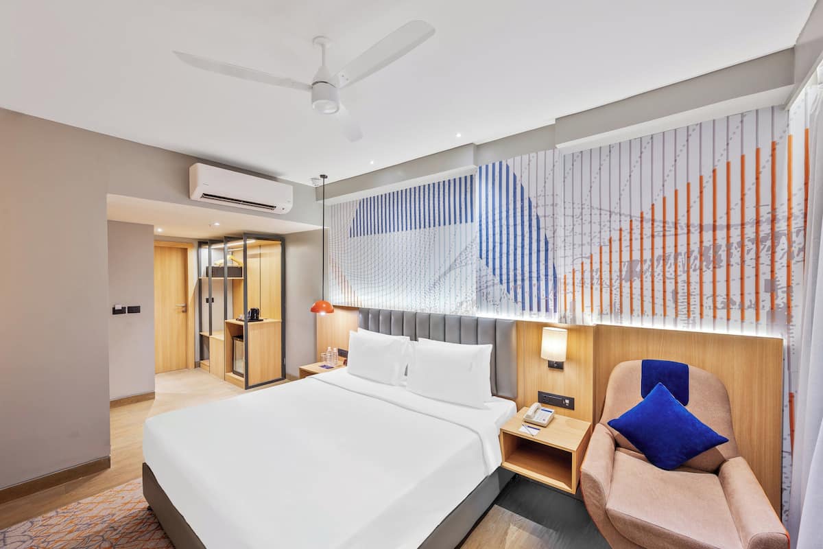 A modern hotel room with a bed, a chair, a desk, and a window with blinds.