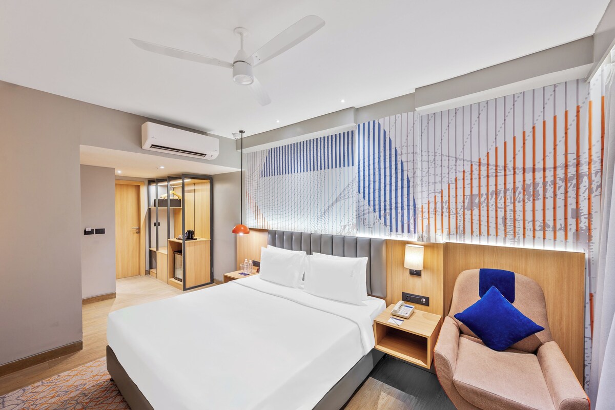 A modern hotel room with a bed, a chair, a desk, and a window with blinds.
