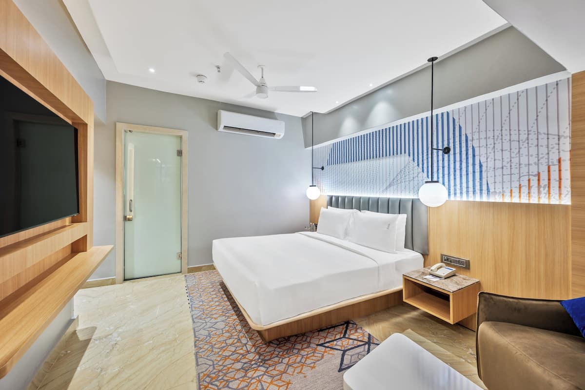 A modern hotel room with a large bed, a TV, a desk with a lamp, and a wall-mounted air conditioner.
