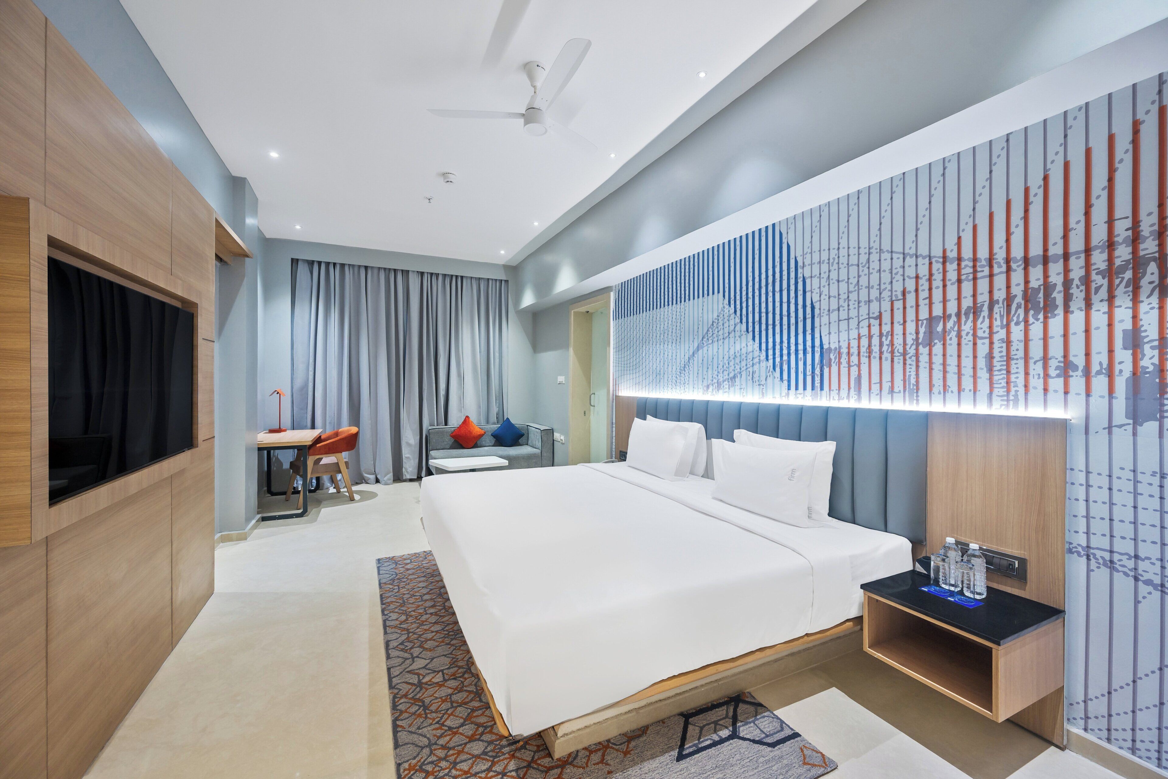 A modern hotel room with a large bed, a desk, a chair, a TV, and a wall-mounted shelf with bottles.