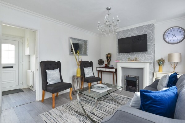 A modern living room with two black armchairs, a glass coffee table, a grey sofa, a fireplace, a mounted TV, and a wall clock. - Suspire House -sleeps6-garden-bbq-coastal Retreat (New Romney)
