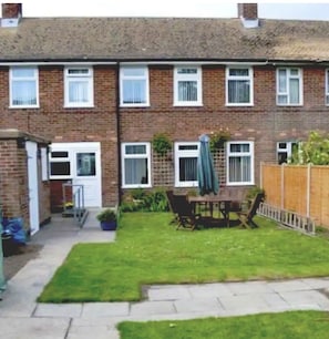 A two-story brick house with a small front garden, a patio with a table and chairs, and a wooden fence.