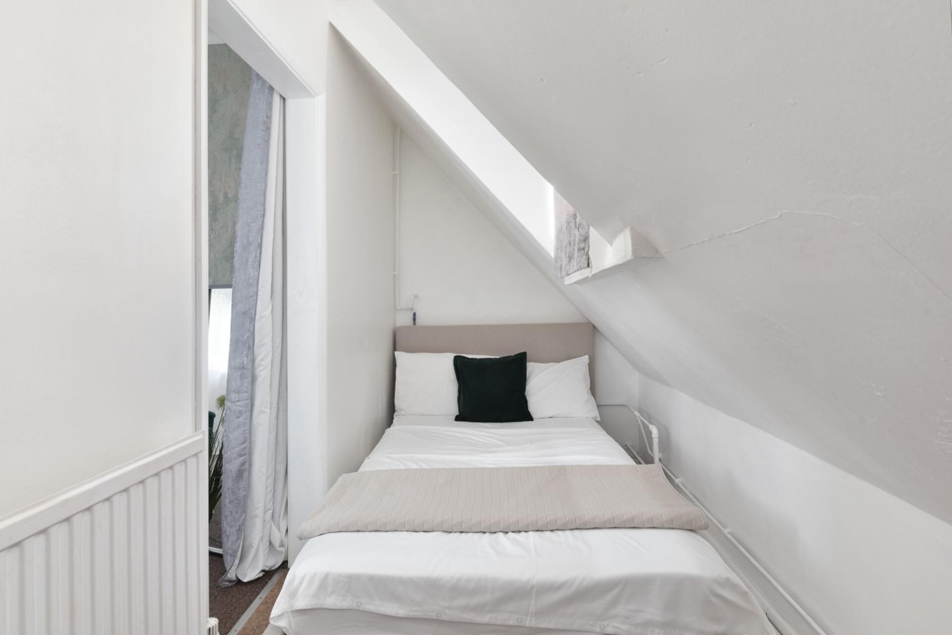 Una habitación estrecha con paredes blancas y techo inclinado, una cama con ropa blanca y almohada negra, y un ventanal pequeño.
