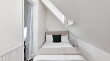 Una habitación estrecha con paredes blancas y techo inclinado, una cama con ropa blanca y almohada negra, y un ventanal pequeño.